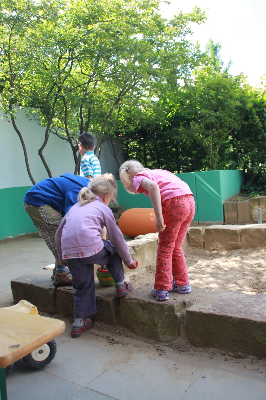Zwei Kinder beugen sich über die Mauer des Sandkastens im Außengelände und graben im Sand.