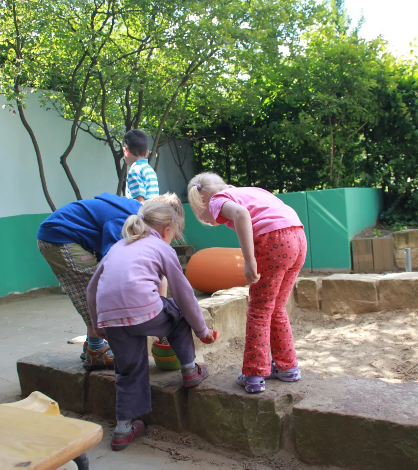 Zwei Kinder beugen sich über die Mauer des Sandkastens im Außengelände und graben im Sand.