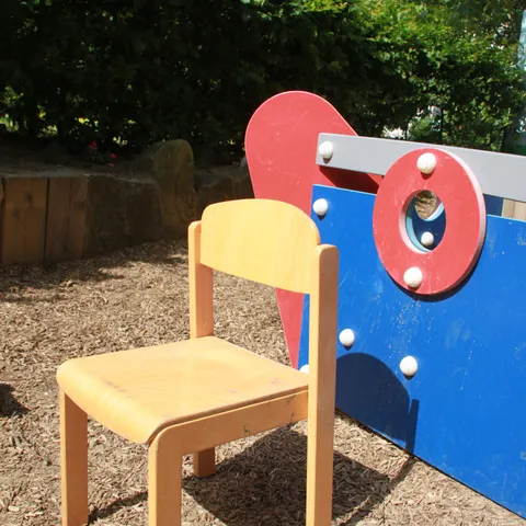 Ein gelber Kinderstuhl steht im Sand vor einem blau-roten Spielhaus.