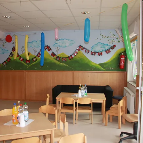 Gruppenraum mit Kindertischen und -stühlen, an der Wand ein buntes Wandbild.