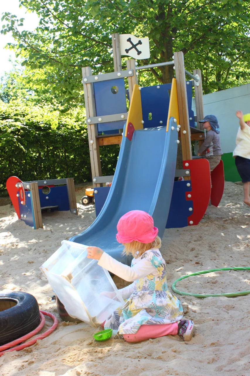 Ein Kind mit Sonnenhut spielt am Sandkasten neben Rutsche und Wasserschlauch.