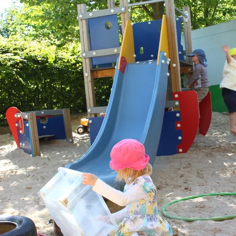 Ein Kind mit Sonnenhut spielt am Sandkasten neben Rutsche und Wasserschlauch.