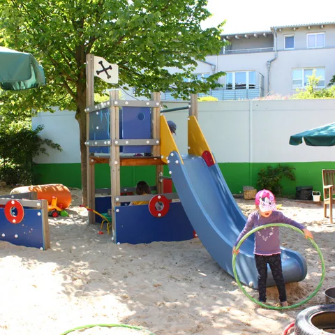 Blick über das Außengelände mit Klettergerüst, Rutsche und Sonnenschirm.