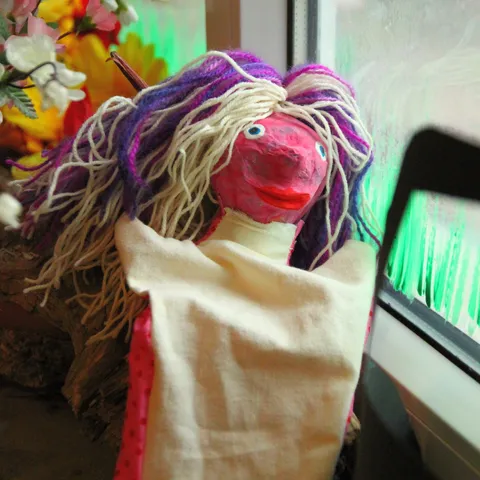 Handpuppe mit gelbem Wollhaar vor einer Fensterbank mit Blumen.