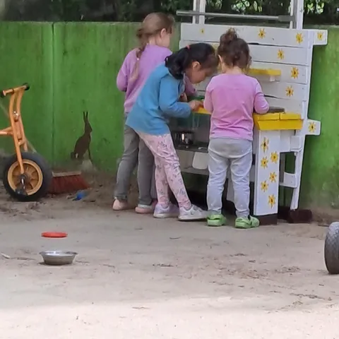 Drei Kinder stehen gemeinsam an der Matschküche, daneben ein Dreirad im Sand.