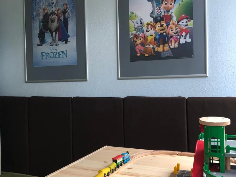 Heller Bauraum mit einem großen Holztisch, auf dem eine Holzeisenbahn mit Zug und Parkhaus aufgebaut ist. An der hellblauen Wand hängen zwei gerahmte Kinderposter.