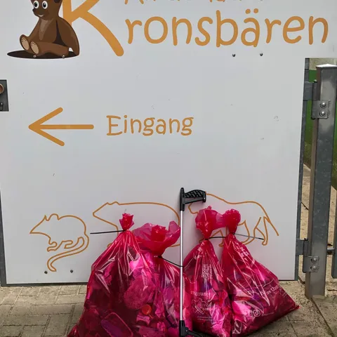 Vier volle rosa Müllsäcke und ein Müllgreifer lehnen am Eingangsschild des Kinderladens.