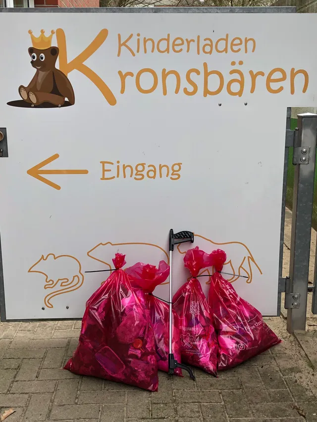 Vier prall gefüllte rosa Müllsäcke und ein Müllgreifer lehnen am Eingangsschild des Kinderladens Kronsbären.