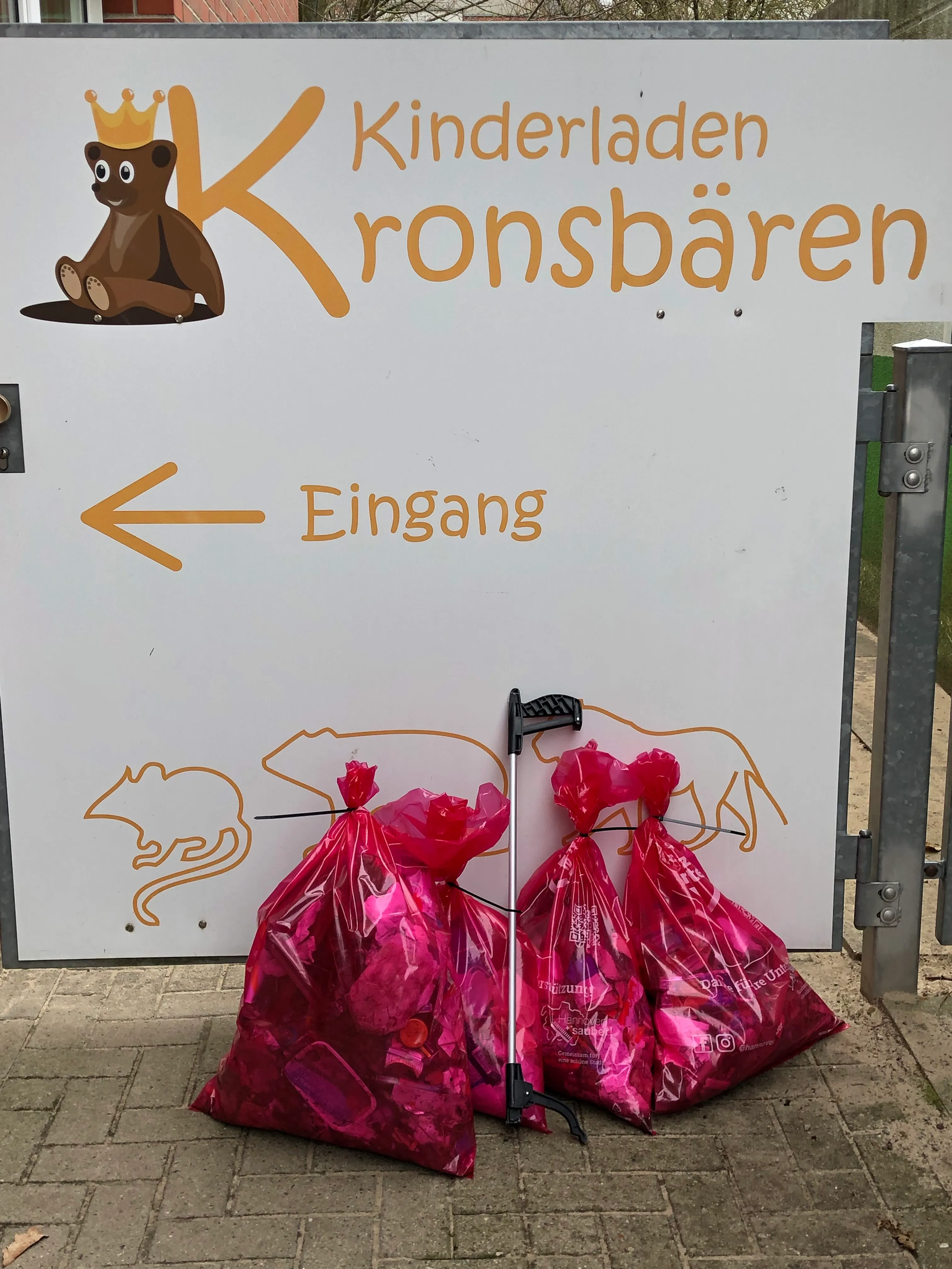 Vier prall gefüllte rosa Müllsäcke und ein Müllgreifer lehnen am Eingangsschild des Kinderladens Kronsbären.