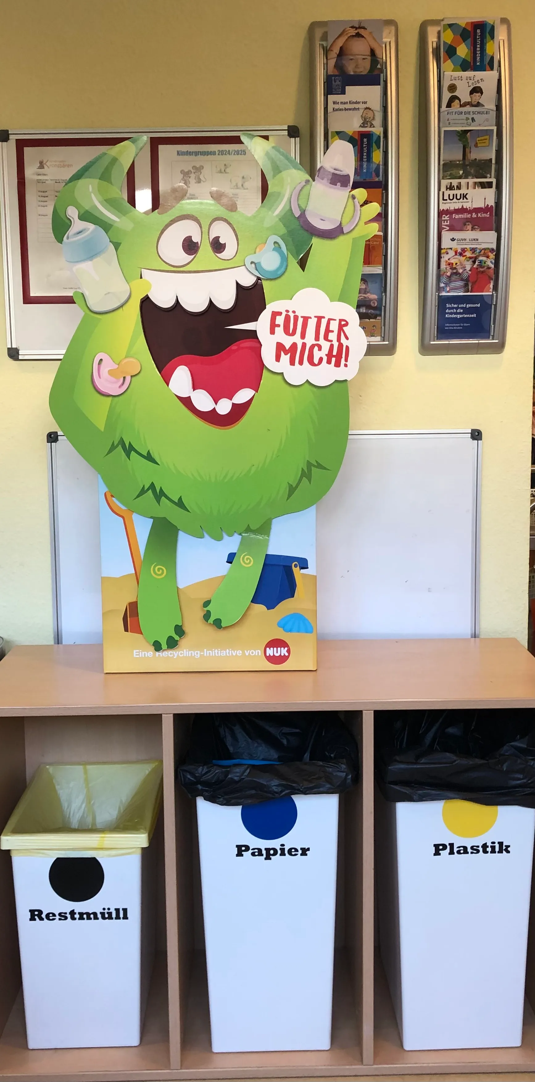 Große Pappfigur eines grünen Monsters mit weit geöffnetem Mund und der Sprechblase „Fütter mich!“, in den Händen Babyflaschen und Schnuller. Darunter stehen drei Sammelbehälter für Restmüll, Papier und Plastik.