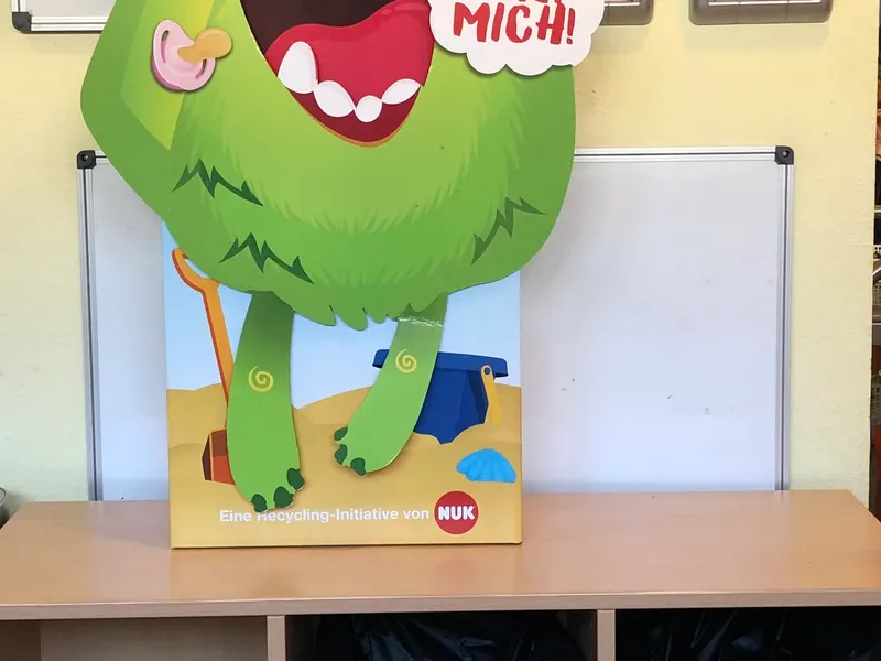 Große Pappfigur eines grünen Monsters mit weit geöffnetem Mund und der Sprechblase „Fütter mich!“, in den Händen Babyflaschen und Schnuller. Darunter stehen drei Sammelbehälter für Restmüll, Papier und Plastik.